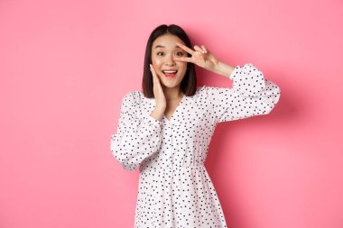 Güzel ve kadınsı Asyalı kadın kawaii işaretini gözlerinde gösteriyor ve kameraya hayret içinde bakıyor, pembe arka planda elbiseler içinde duruyor.