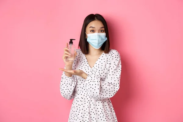 Covid-19, conceito de pandemia e estilo de vida. Mulher coreana bonita em vestido e máscara médica mostrando higienizador das mãos, recomendando anti-séptico, de pé sobre fundo rosa — Fotografia de Stock