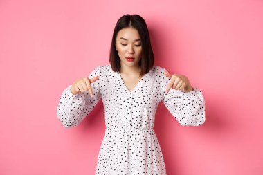 Romantik Asyalı kadın dikizliyor, parmakları dipte, ürün indirimine bakıyor, pembe arka planda duruyor.
