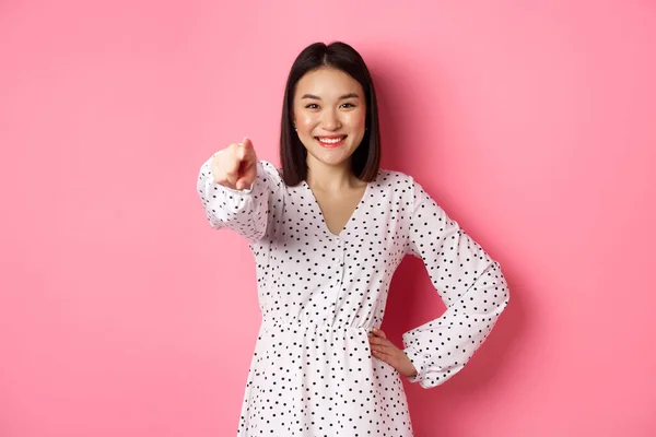 Confiante mulher asiática em vestido apontando para você, sorrindo e olhando para a câmera, de pé contra o fundo rosa — Fotografia de Stock