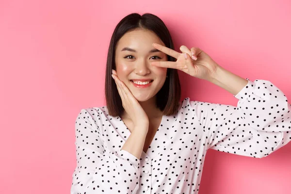 Concept de beauté et style de vie. Gros plan de mignon asiatique femme montrant signe de paix et toucher la joue, souriant heureux à la caméra, debout sur fond rose — Photo