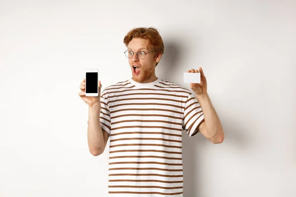 Conceito de compras e finanças. Jovem incrível com cabelo vermelho mostrando cartão de crédito de plástico e tela em branco do smartphone, olhando para a tela impressionado, fundo branco — Fotografia de Stock