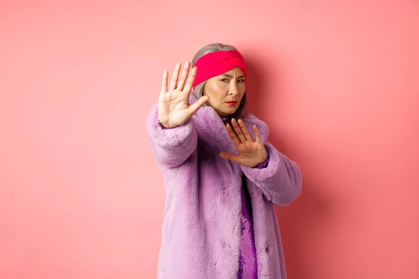 Ασιάτης ανώτερος θηλυκό μοντέλο σε κομψό μοβ παλτό χειμώνα στέκεται στο θύμα θέτουν, επέκταση χέρι σε στάση χειρονομία και παρακαλώντας για έλεος, στέκεται σε ροζ φόντο — Φωτογραφία Αρχείου