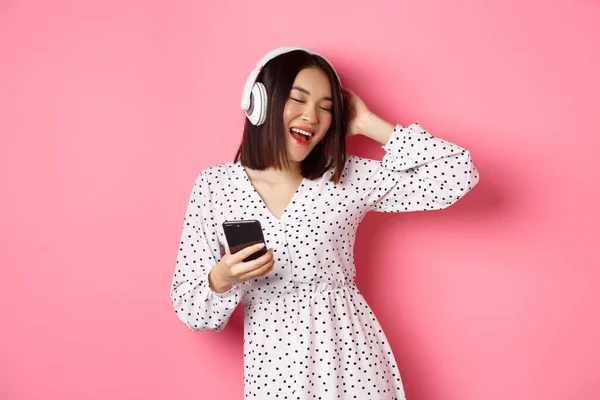 Beztroska azjatycka kobieta bawiąca się, tańcząca i słuchająca muzyki w słuchawkach, trzymająca telefon komórkowy, stojąca w sukience na różowym tle — Zdjęcie stockowe