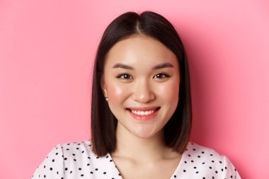 Beauty and lifestyle concept. Headshot of beautiful asian woman smiling, looking at camera happy and romantic, standing against pink background clipart