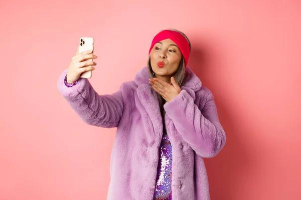 Modekonzept. Schöne und trendige asiatische reife Frau im Kunstpelzmantel mit Partykleid, macht Selfie, schickt Küsse in die Smartphone-Kamera, posiert vor rosa Hintergrund — Stockfoto