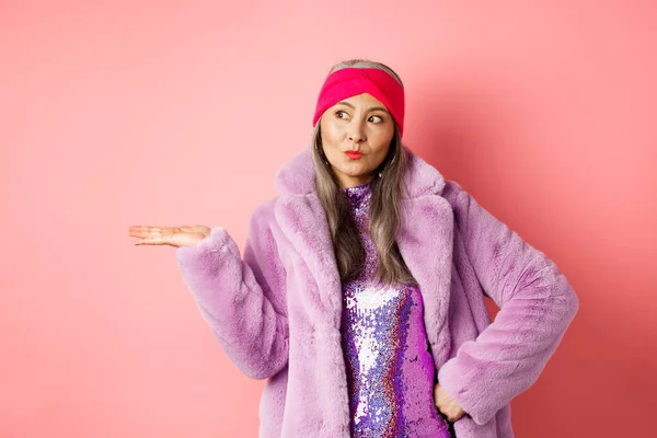 Mode- und Einkaufskonzept. Stilvolle asiatische Seniorin im lila Winteroutfit demonstriert Produkt auf der Hand. Coole alte Dame in trendiger Kleidung mit etwas auf der Handfläche, rosa Hintergrund — Stockfoto