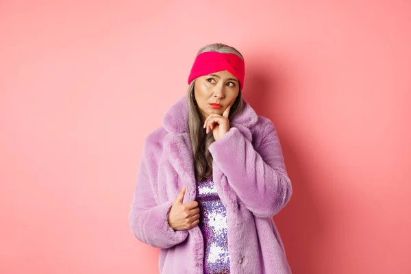 Moda asiática mulher sênior olhando duvidoso, pensando e franzindo a testa indeciso, de pé em casaco de pele falso roxo e olhando para a esquerda, de pé sobre fundo rosa — Fotografia de Stock