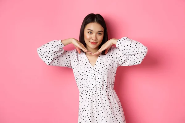 Hermosa mujer asiática en vestido de moda haciendo cara linda, cogida de la mano cerca de la mandíbula y mirando coqueta a la cámara, de pie sobre fondo rosa —  Fotos de Stock