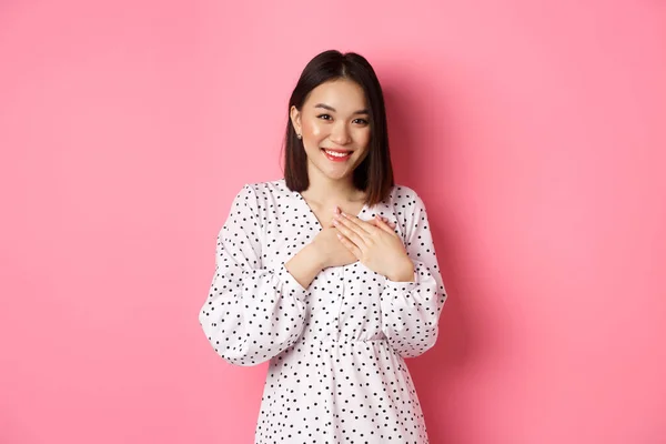 Chica coreana agradecida en vestido sonriente, cogida de la mano en el corazón y mirando agradecida a la cámara, tocada con gesto agradable, de pie sobre fondo rosa —  Fotos de Stock