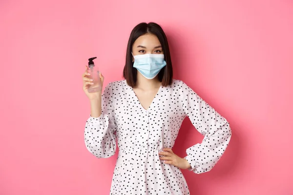 Covid-19, pandemia ja elämäntapa käsite. Kaunis korealainen nainen mekko ja lääketieteellinen naamio osoittaa käsi sanitizer, suositella antiseptinen, seisoo vaaleanpunainen tausta — kuvapankkivalokuva