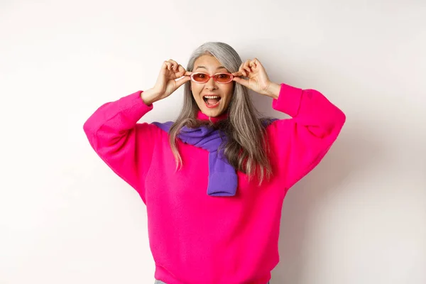 Модная азиатская старшая женщина пробует новые солнцезащитные очки, смотрит изумленно и весело на камеру, стоя в модном свитере на белом фоне — стоковое фото