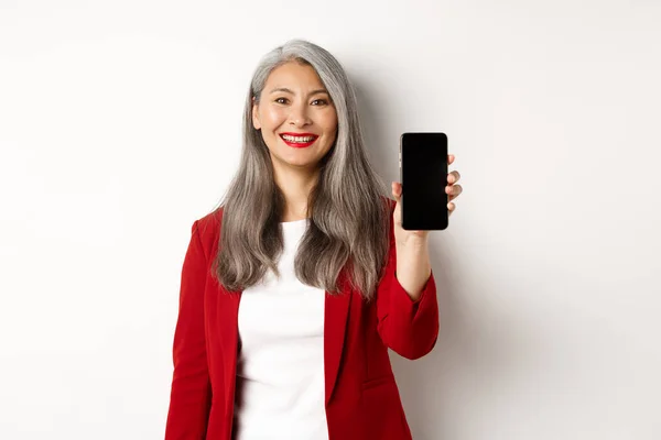 Äldre affärskvinna i röd blazer, visar tom smartphone skärm och leende, demonstrera mobilapp, vit bakgrund — Stockfoto