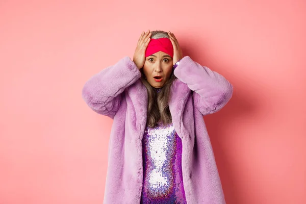 Schockiert und alarmiert steht eine asiatische Seniorin in lila Kunstpelzmantel vor rosa Hintergrund, hält die Hände auf dem Kopf und gerät in Panik. — Stockfoto