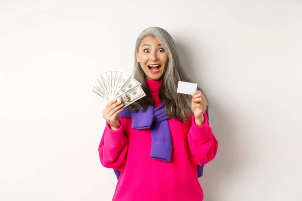 Shopping concept. Amazed asian granny showing plastic credit card and money dollars, want to buy something, standing over white background — Stock Photo, Image