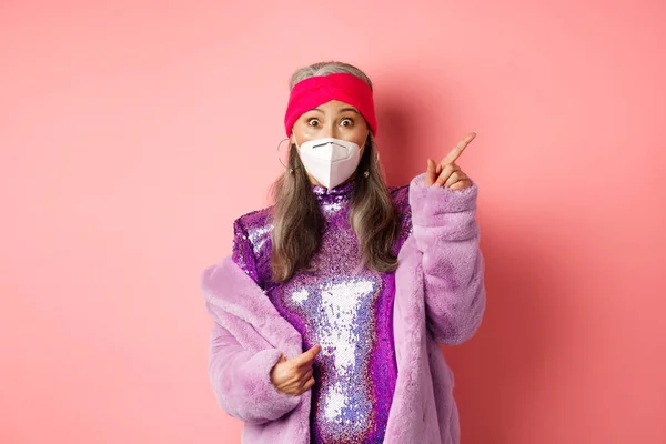 Coronavirus και ιδέα για ψώνια. Εκπληκτική Ασιάτισσα ηλικιωμένη με μάσκα προσώπου να κοιτάζει την κάμερα, να δείχνει το διαφημιστικό της πάνω δεξιάς γωνίας, να στέκεται πάνω από ροζ φόντο — Φωτογραφία Αρχείου