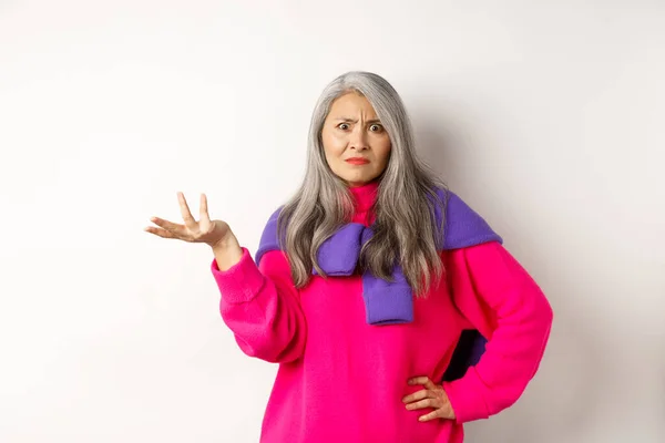 Wütend und verwirrt breitete eine asiatische Seniorin ihre Hand zur Seite aus und starrte verdutzt in die Kamera, die im rosafarbenen Pullover vor weißem Hintergrund stand. — Stockfoto