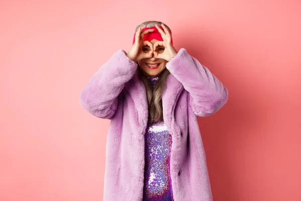 Mode und Shopping. Glückliche asiatische Großmutter in stylischem Faur-Pelzmantel, schaut durch eine Handbrille, macht Ferngläser und lächelt in die Kamera, steht über rosa Hintergrund — Stockfoto