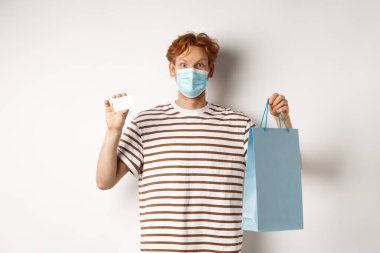 Covid-19 ve yaşam tarzı kavramı. Kızıl saçlı neşeli genç adam, tıbbi maske takıyor, dükkandan alışveriş torbası ve plastik kredi kartı gösteriyor.