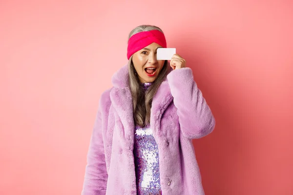 Nákupní a módní koncept. Krásná asijské středního věku žena ukazující plastové kreditní karty na obličeji a vypadá nadšeně na kameru, stojící nad růžovým pozadím — Stock fotografie