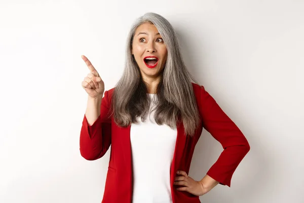 Överraskad senior asiatisk dam pekar övre vänstra hörnet, kolla promo erbjudande och leende förvånad, står i röd blazer över vit bakgrund — Stockfoto