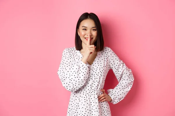 Όμορφη Κορεάτισσα γυναίκα σε μοντέρνο φόρεμα ζητώντας να κρατήσει μυστικό, hushing με απαλό χαμόγελο, σχεδιάζει μια έκπληξη, στέκεται πάνω από ροζ φόντο — Φωτογραφία Αρχείου
