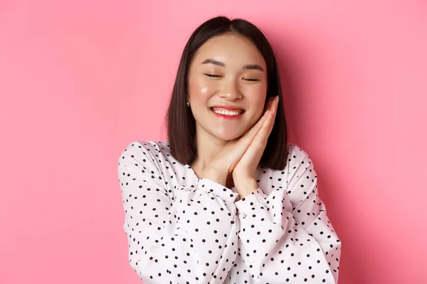 Conceito de beleza e estilo de vida. Close-up de bela e sonhadora mulher asiática dormindo em suas mãos, fechar os olhos e sorrindo, sonhando acordado em fundo rosa — Fotografia de Stock