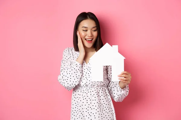 Immobiliare. Carino donna asiatica in cerca di appartamento, guardando felice a modello di casa di carta, in piedi sullo sfondo rosa — Foto Stock