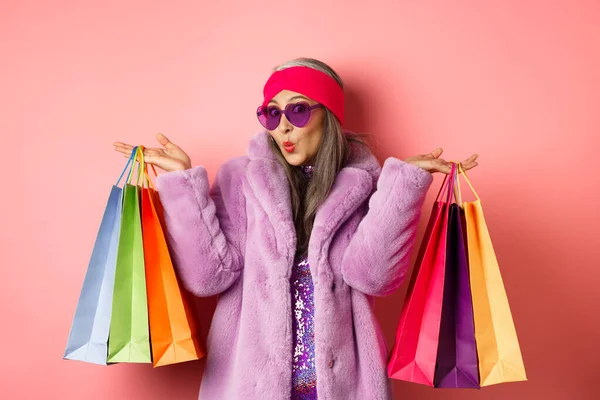 Stilvolle asiatische Seniorin geht einkaufen, trägt trendige Kleidung und Sonnenbrille, hält Einkaufstaschen mit Kleidung und Einkäufen in der Hand, rosa Hintergrund — Stockfoto