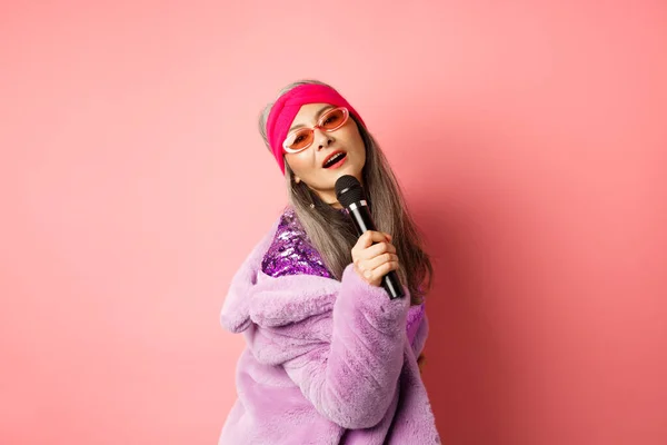 Κομψή και αυθάδης ώριμη γυναίκα από την Ασία ερμηνεύει τραγούδι στη σκηνή, κρατώντας μικρόφωνο και τραγουδώντας καραόκε, φορώντας μοντέρνα γυαλιά ηλίου σε σχήμα καρδιάς και faux γούνα παλτό, ροζ φόντο — Φωτογραφία Αρχείου