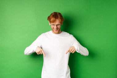 Gözlüklü ve uzun kollu yakışıklı kızıl adam reklamlara bakıyor, parmaklarıyla işaret ediyor ve gülümsüyor, yeşil arka planda dikiliyor.