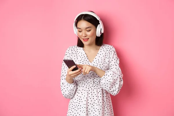 Piękna Azjatka SMS-uje na smartfonie, słucha muzyki w słuchawkach, stoi nad różowym tłem — Zdjęcie stockowe