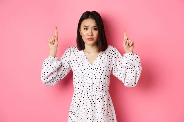 Molesto chica asiática señalando los dedos hacia arriba, mostrando pancarta promocional, mirando fijamente a la cámara con mirada triste decepcionado, de pie en el vestido sobre fondo rosa —  Fotos de Stock
