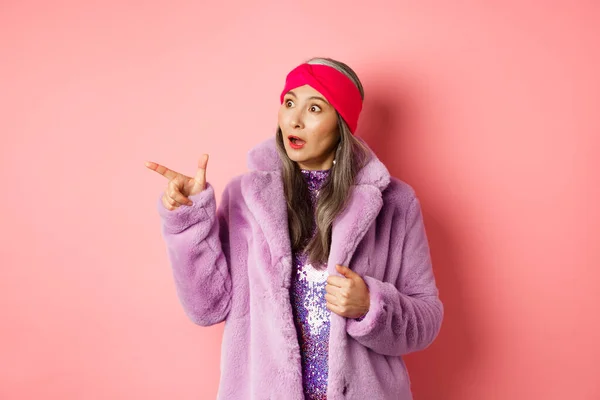 Fashion and shopping concept. Trendy elderly asian female in fake fur and glittering dress pointing, looking left with amazement, checking out offer, pink background — Stock Photo, Image