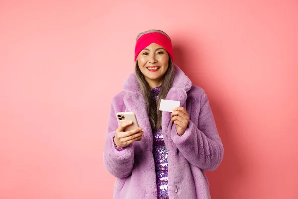 Online-Shopping und Modekonzept. Lächelnde Frau mittleren Alters in stylischem Pelzmantel mit Handy und Plastikkreditkarte, glücklich stehend auf rosa Hintergrund — Stockfoto
