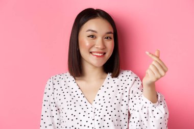 Güzellik ve yaşam tarzı konsepti. Sevgililer gününde kalp işareti gösteren, gülümseyen ve romantik hisseden, pembe arka planda duran güzel Asyalı kadının yakın çekimi.