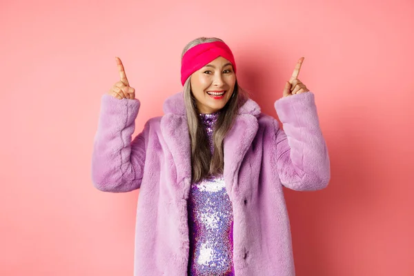 Elegante mulher asiática sorrindo feliz, apontando os dedos para cima e mostrando propaganda, vestindo inverno casaco de pele falsa e headband, fundo rosa — Fotografia de Stock
