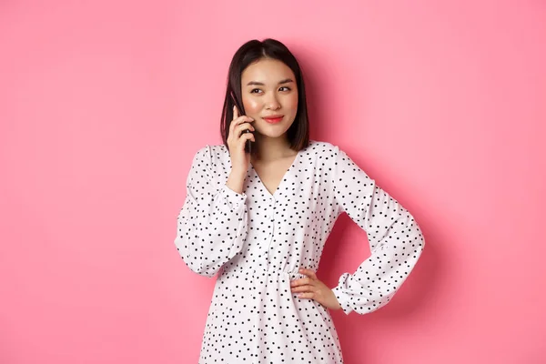 Mujer coreana atractiva haciendo una llamada telefónica, sosteniendo el teléfono inteligente cerca de la oreja y sonriendo, de pie sobre el fondo rosa —  Fotos de Stock