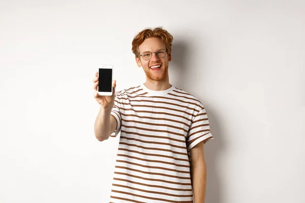 技术和电子商务概念。戴眼镜快乐的红头发年轻人，脸上挂着空白的智能手机屏幕，满意地微笑着，站在白色的背景上 — 图库照片