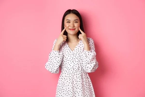 Χαριτωμένο κοκκινισμένο κορίτσι από την Ασία να τσιμπάει μάγουλα, χαμογελώντας χαρούμενο στην κάμερα, φορώντας ρομαντικό λευκό φόρεμα, στέκεται πάνω από ροζ φόντο — Φωτογραφία Αρχείου