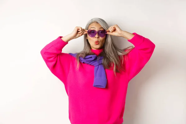 Modische asiatische Seniorin in Sonnenbrille und stylischem rosa Pullover Puckerlippen, zieht die Augenbrauen hoch und schaut überrascht in die Kamera, weißer Hintergrund — Stockfoto