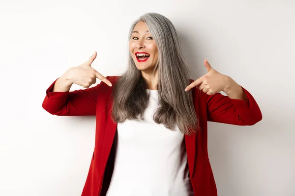 Ehrgeizige Seniorin zeigt mit dem Finger in die Mitte, zeigt Logo und lächelt, steht über weißem Hintergrund — Stockfoto