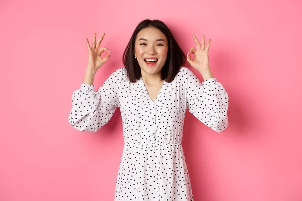 Bastante joven asiático mujer en vestido mostrando ok signo, alabando y mostrando aprobación, buscando satisfecho, de pie sobre rosa fondo —  Fotos de Stock