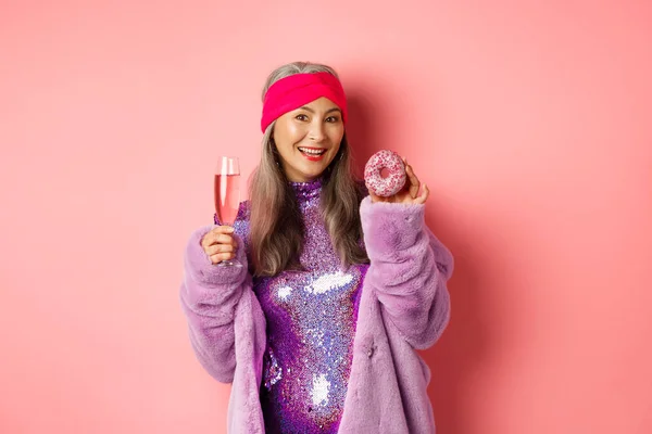 Bella ed elegante asiatica anziana che si diverte, beve champagne e mangia ciambelle dolci, sorride alla macchina fotografica, in piedi su sfondo rosa — Foto Stock