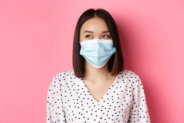Covid-19, conceito de pandemia e estilo de vida. Bonita asiática modelo feminino em máscara médica rindo, sorrindo e olhando para a esquerda no espaço de cópia, de pé sobre fundo rosa — Fotografia de Stock