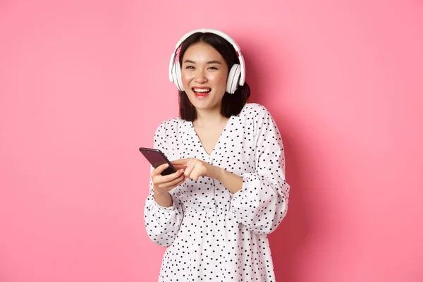 Piękna Azjatka słuchająca muzyki w słuchawkach, korzystająca z telefonu komórkowego, uśmiechnięta szczęśliwa przed kamerą, stojąca nad różowym tłem — Zdjęcie stockowe
