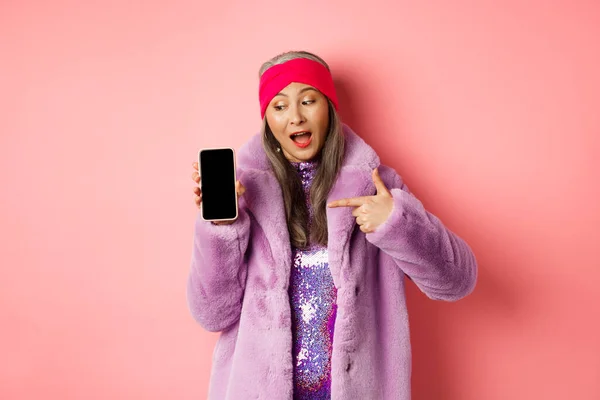 Online-Shopping und Modekonzept. Coole asiatische Oma im stylischen Kunstpelzmantel zeigt mit dem Finger links auf leeren Smartphone-Bildschirm, checkt Internet-Promo, rosa Hintergrund — Stockfoto