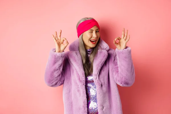 Fashion and shopping concept. Stylish asian granny saying all okay and winking. Trendy senior woman in winter fake-fur coat show OK sign, praising good product, pink background — Stock Photo, Image