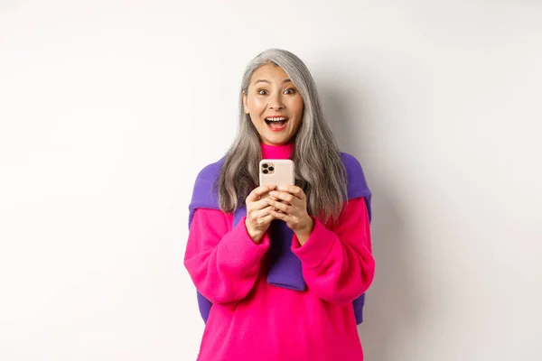 Zaskoczona azjatycka kobieta uśmiechnięta do kamery po przeczytaniu promocji na smartfonie, stojąca z telefonem komórkowym na białym tle — Zdjęcie stockowe