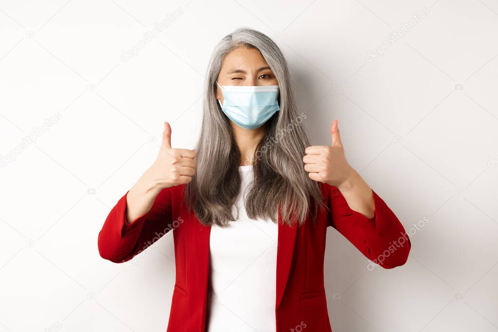 Coronavirus and business concept. Asian female entrepreneur in face mask looking cheerful, showing thumb-up in approval, white background